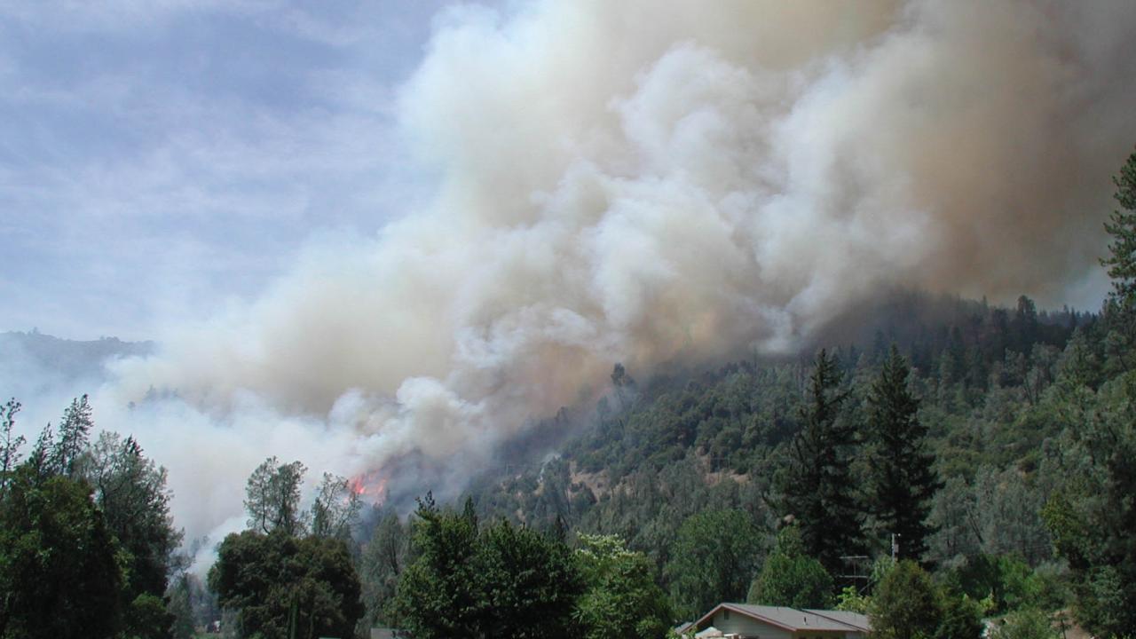 Smoke from wildfire billows over hillside with trees and rural home in foreground