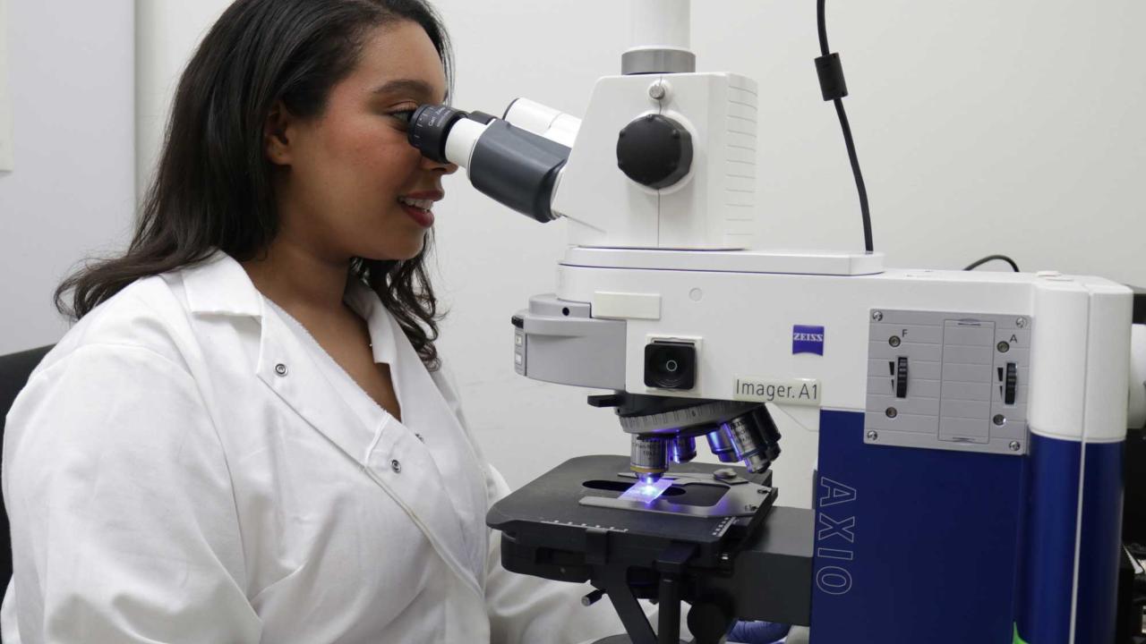 dark-complected woman with long hair in lab coat looking through microscope