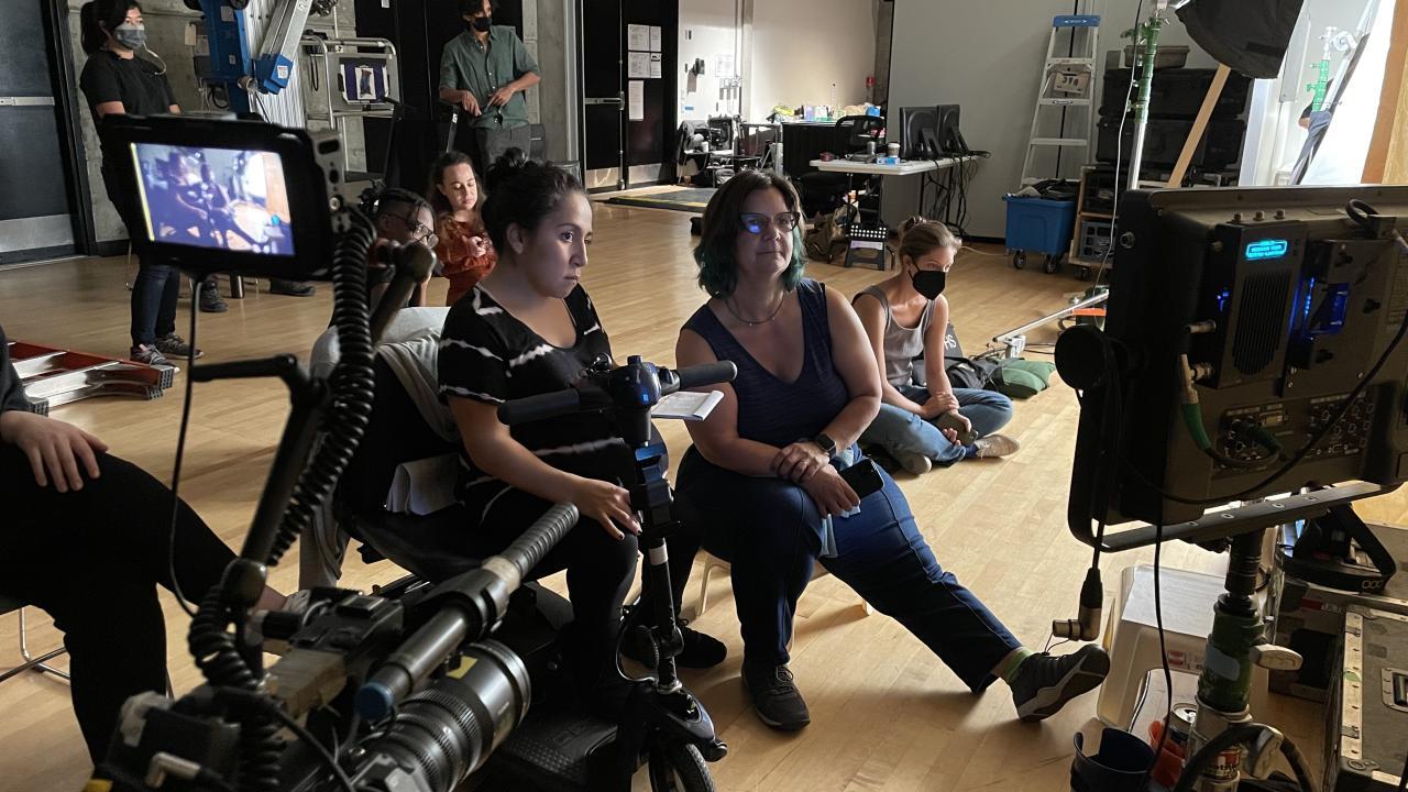 Photo of a set from behind the scenes with two women looking directly at a monitor with other masked individuals in the background.