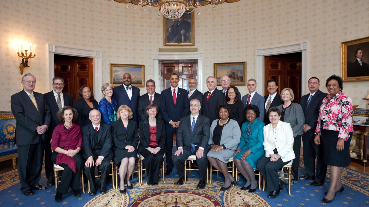 Posed picture of formally dressed men and women in the White House Oval Office. President Barack Obama at center, back row. 