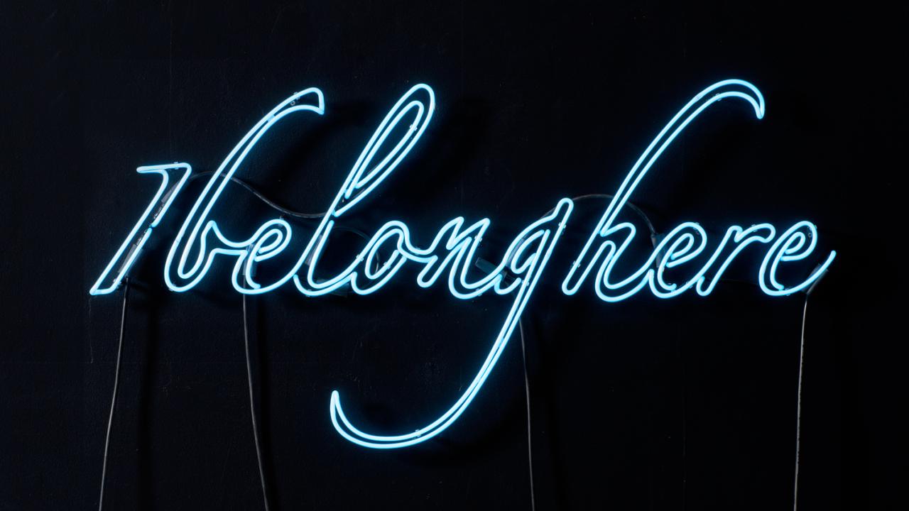 Blue neon lights reading "I belong here" against black text.