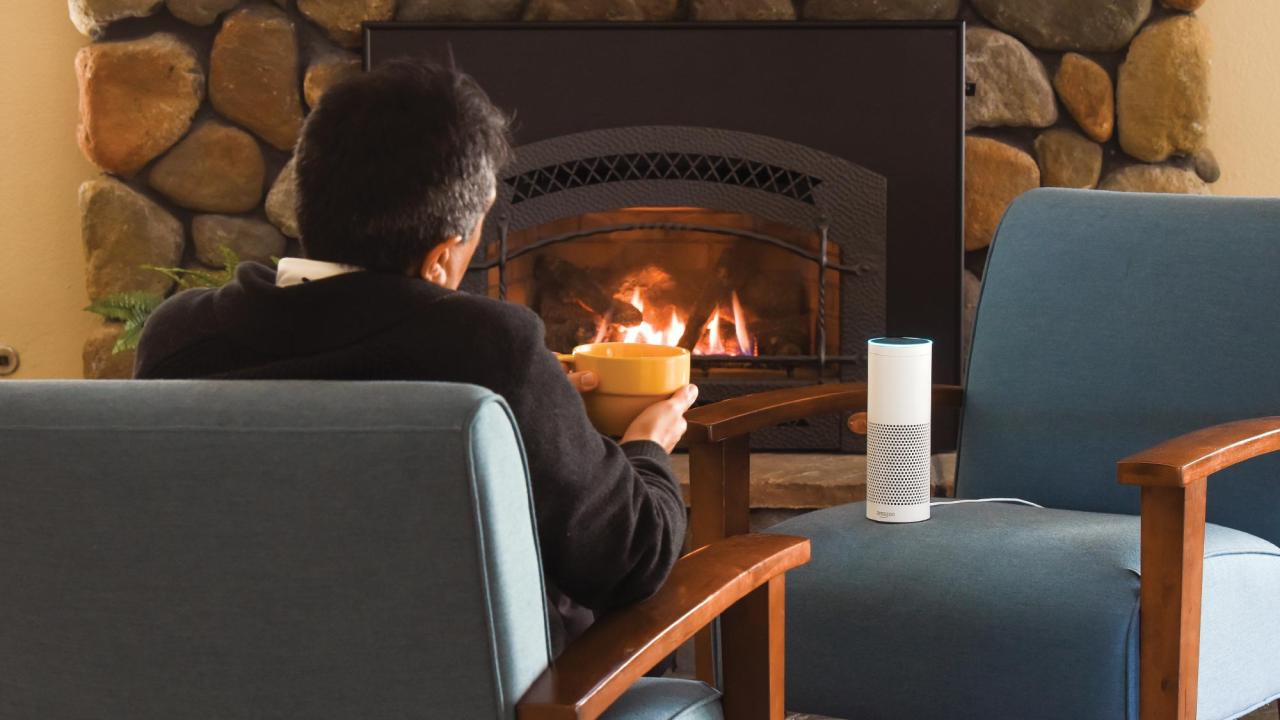 A man sits on a chair in front of a fireplace, back to the camera. A chair faces him with an Amazon Echo device sitting on it, lit up.