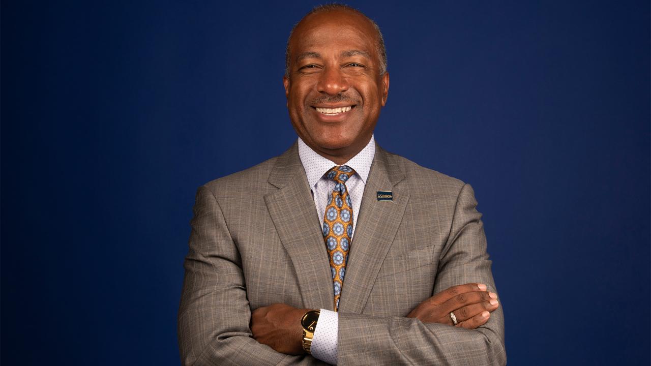 Portrait of Chancellor Gary S. May, standing and smiling with arms crossed in front of a blue background.