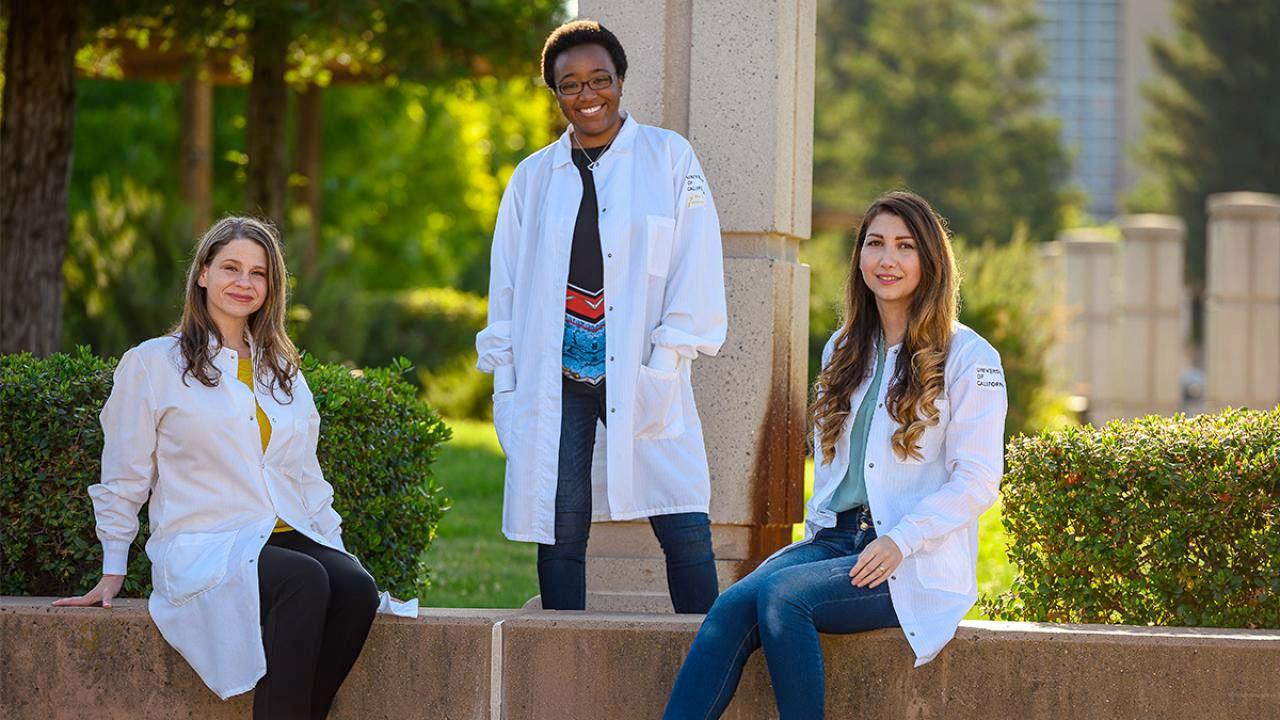 Three female students in lab coats pose for a shot on the UC Davis campus