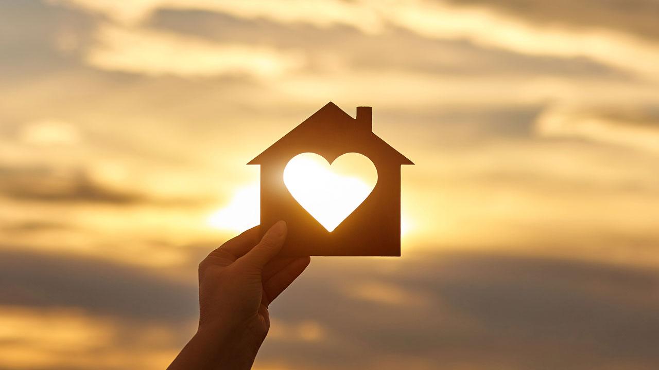 A hand holding up a house cutout with a heart in the middle against a sunset background