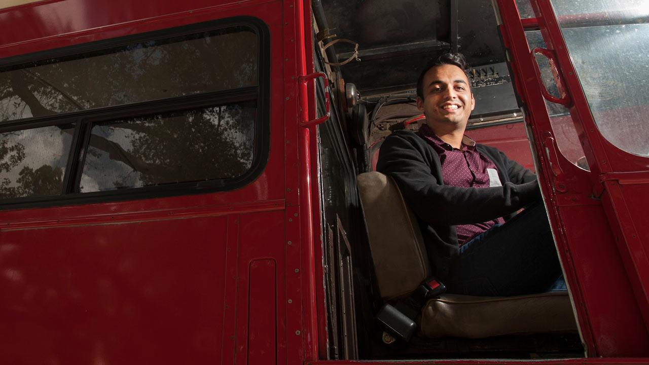 A male student sitting in the driver's seat of a double decker bus.