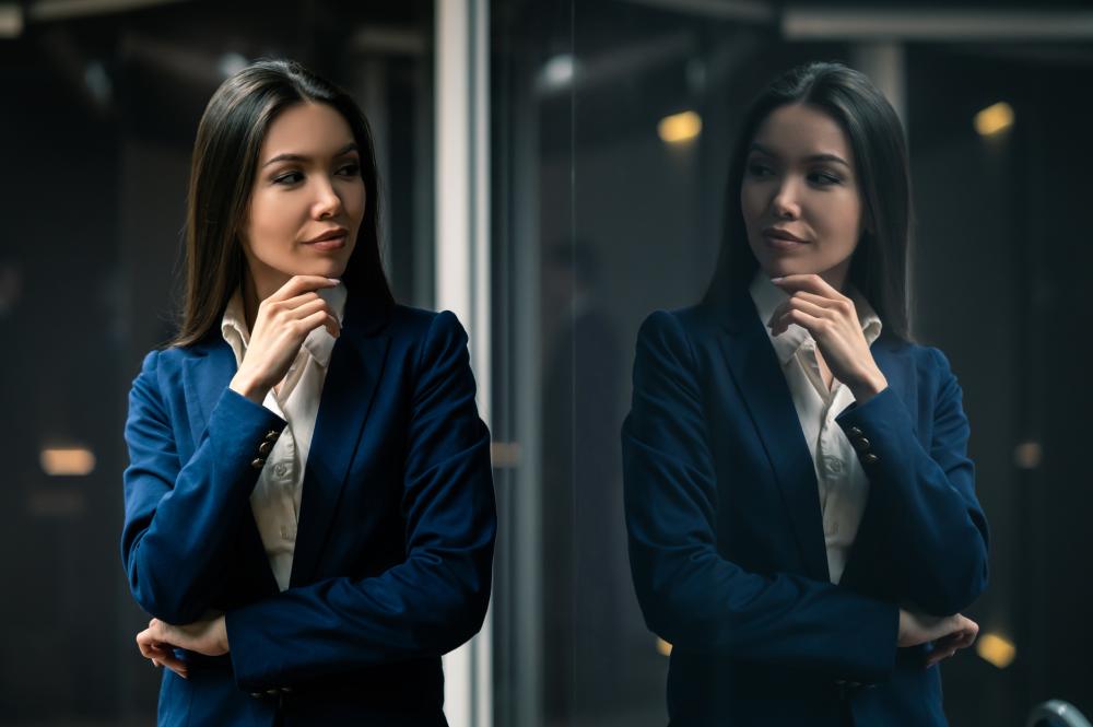 A woman with shoulder length black hair wearing a blue jacket looks sideways at her reflection. 