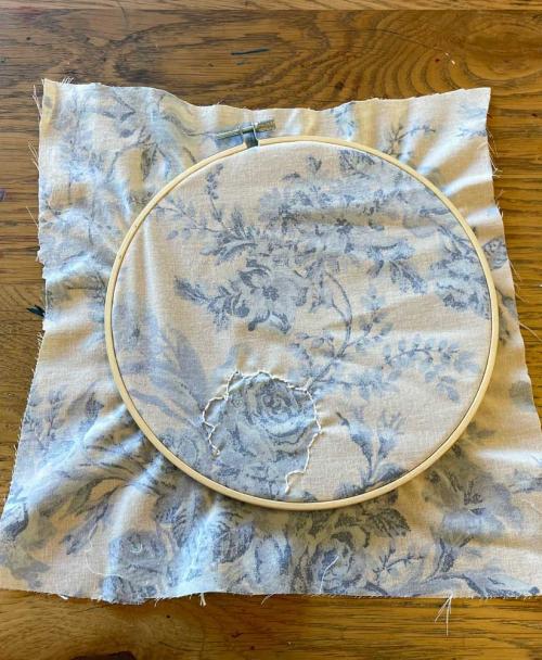 An embroidery hoop holds white fabric with blue flowers. There is white embroidery thread stitched over the outline of one of the flowers.