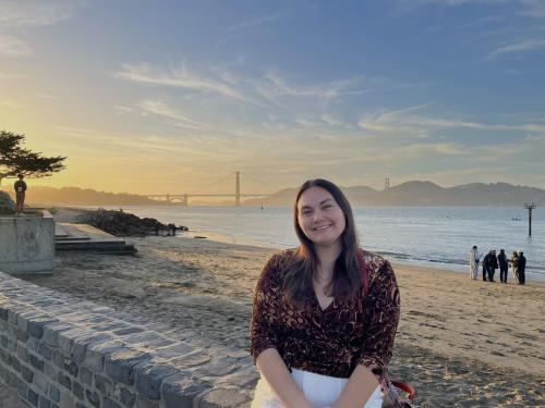 Girl in a brown patterned shirt and white pants sits down in front of a view of the Golden Gate bridge in San Francisco. 