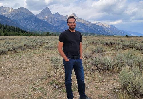 A smiling man wearing a black t-shirt and jeans stands casually against a background of sagebrush and mountains.