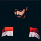 Jai Wolf looking downward, wearing mostly black and standing in front of a black background.