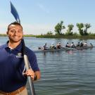 First-generation college student Ricky Zapardiel stands in front of a lake and a rowboat while holding a paddle. 