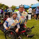 Man in bike helmet and jersey kneels next to a man seated on a recumbent bike with people and event tents behind them