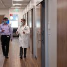 Two men (each with a stethoscope around his neck) chat as they walk in hallways, toward camera.