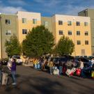 Families gather around cars, in parking lot, unloading students' belongings, with residence hall in background.