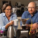 Professors Tonya Kuhl and Bill Ristenpart sit side by side facing us from behind the counter of a lab station. Kulfis is holding a cup of coffee and Ristenpart is sitting with crossed arms on the table, while an instrument is attached to a coffee filter with a yellow cord as the coffee drips into a clear glass cup. Various coffee makers and lab instruments are in front of and behind them.