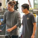 A male student wearing a grey t-shirt and clear protective glasses operates machinery in a student workship, while a male student wearing a blue t-shirt and clear protective goggles stands to the right and looks onward at the machinery.  