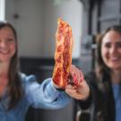 Two women hold up a cultivated meat item that looks like bacon