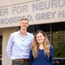 A tall man with blonde hair wearing a light blue shirt stands next to a shorter woman with long brown hair in a dark blue shirt in front of a building entrance. Signage reads Center for Neuroscience Robert D. Grey Hall