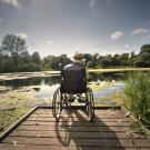 Man in wheelchair on bridge facing water body with back to camera.