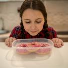 A child stands in a kitchen facing the camera, looking down into a tub of three pink glazed donuts.