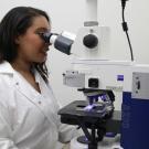 dark-complected woman with long hair in lab coat looking through microscope