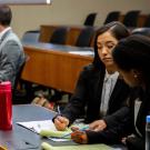 Two students in business attire sit at a desk. They deliberate together over a yellow legal pad.