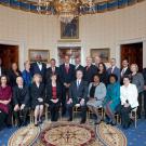 Posed picture of formally dressed men and women in the White House Oval Office. President Barack Obama at center, back row. 