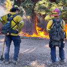 two firefighters stand looking at a wildfire flame