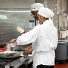 A chef oversees two people cooking at a dining facility stove.