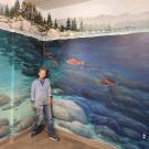 Susie Alexander stands in front of her mural, which features painted rocks, trees, and mountains, as well as rocks and fish in blue water.