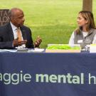 UC Davis Chancellor Gary S. May in suit, sits at table, chatting with two Aggie Mental Health Ambassadors are tabling, on the Quad