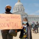 A woman and child with the San Francisco city council building in the background. Both wear facemarks. The woman holds a cardboard sign reading "Domestic Workers are Essential #SB321"