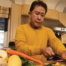 A man in a yellow shirt in his home kitchen slices vegetables.