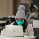 Wiman in mask and knitted cap, at laptop, in Memorial Union