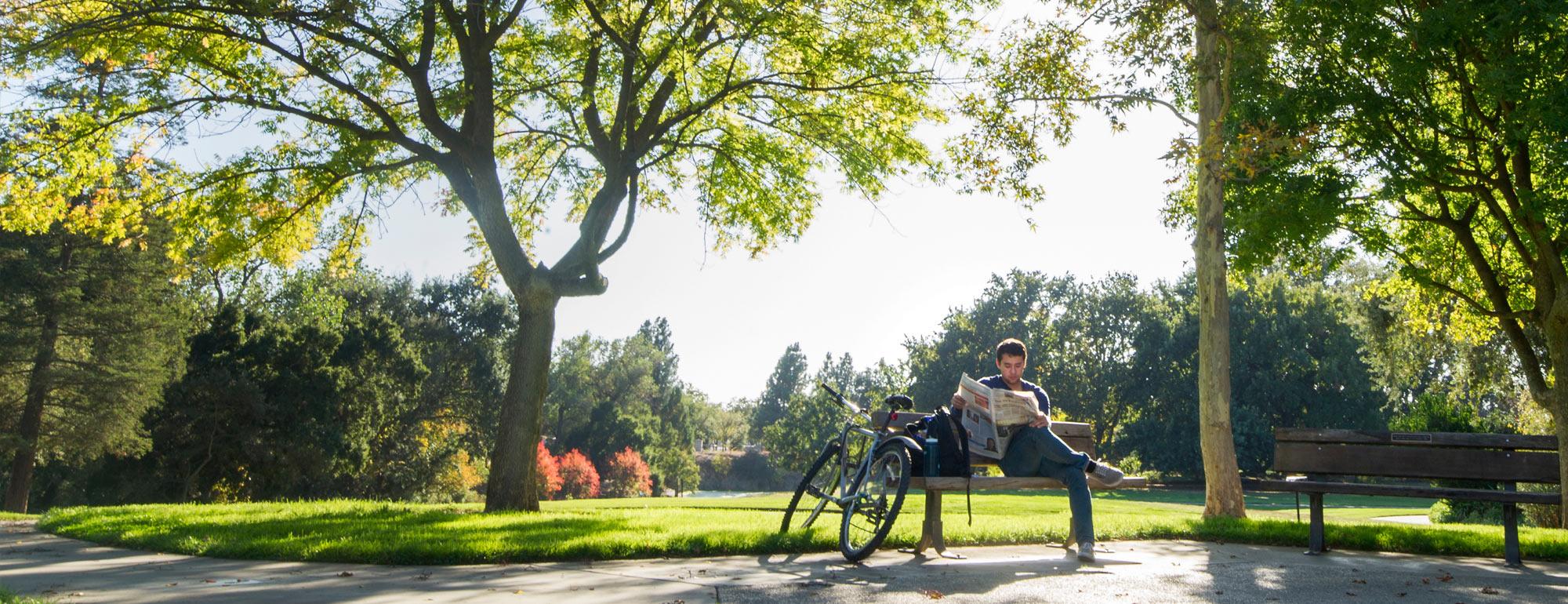 A UC Davis student sits on a bench reading a newspaper next to his bike in front of the beautiful trees of the UC Davis arboretum
