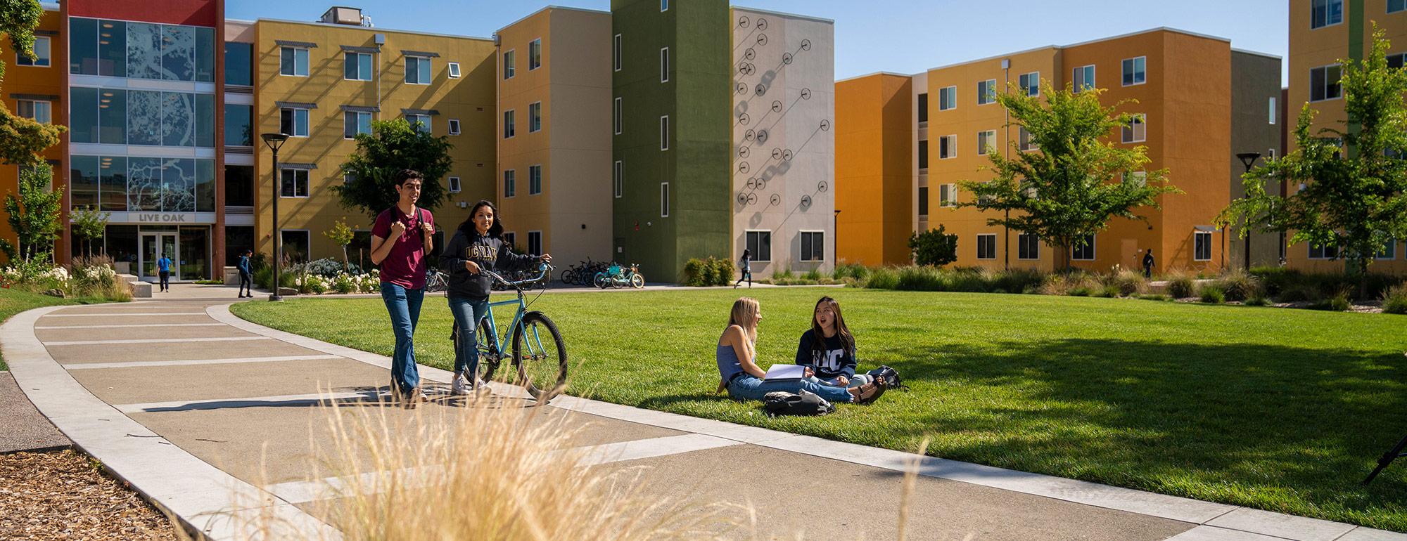 two student walking a bike while two more students are sitting on the grass in front of the residence halls