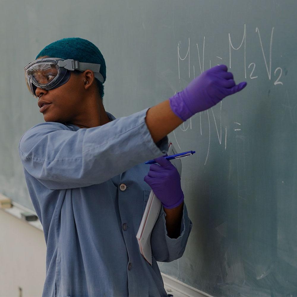 A UC Davis graduate student teaching assistant with short blue hair and wearing a lab coat points to a chemical equation on a blackboard.