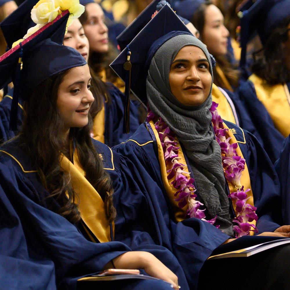 A close up of two students in graduate cap and gown in a group of students attending a graduation ceremony.
