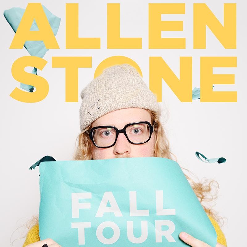 Allen Stone wearing a wool cap and black-framed glasses, holding up a aqua-marine sheet that says "Fall Tour."