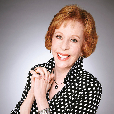 A portrait photo of Carol Burnett smiling, hands folded, wearing a black blouse with white polka-dots and popped collar.