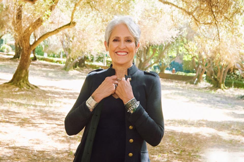 Joan Baez wearing a Nehru-style jacket, smiling and standing outside with old-growth trees in the background.