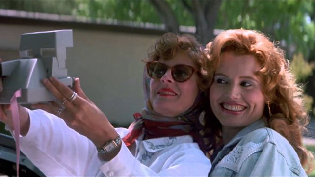 Thelma and Louise taking a selfie in their car with a Polaroid camera.