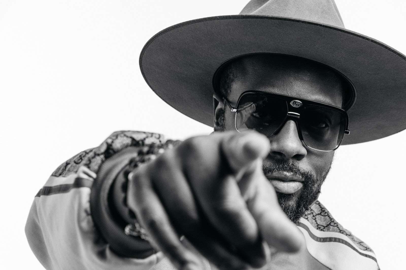 Wyclef Jean wearing a wide-brim hat and sunglasses and pointing into the camera.